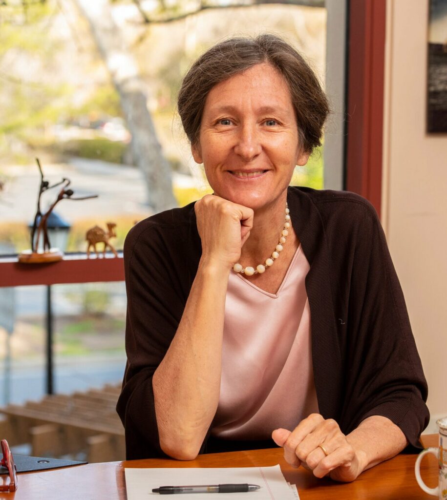 Le docteur Anna Lembke assise à un bureau, s'appuyant sur sa main avec un léger sourire, portant un chemisier rose, un cardigan foncé et un collier de perles, dans une pièce avec une grande fenêtre et quelques petites sculptures visibles en arrière-plan.